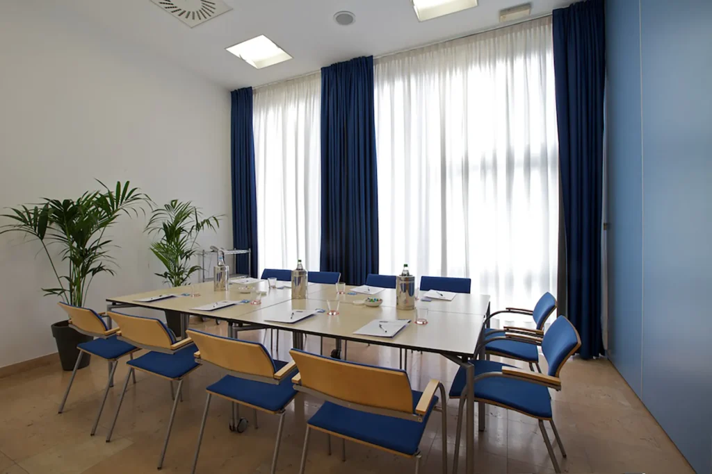 Nell'immagine, la Sala Meeting Vescovo dell'Hotel Fiera Verona: si vede un tavolo unico circondato da sedie: davanti a ogni sedia c'è un blocco di carta con una penna e un bicchiere di vetro. Al centro del tavolo, tre bottiglie d'acqua.