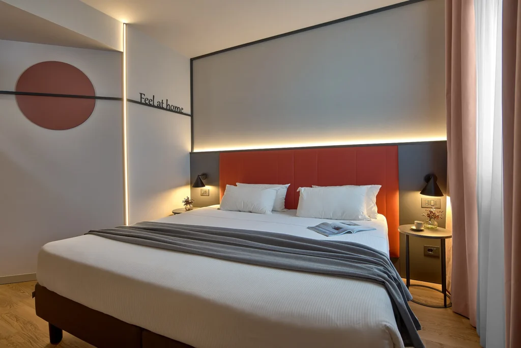 Nell'immagine, la camera Superior dell'Hotel Fiera Verona: si vede il letto matrimoniale e, di fianco, la scritta "Feel at Home" sulla parete.